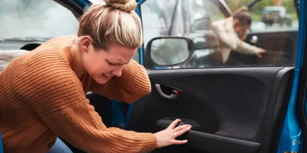 A woman is getting out of her car after a car accident. She is holding the back of her neck after suffering whiplash injuries.