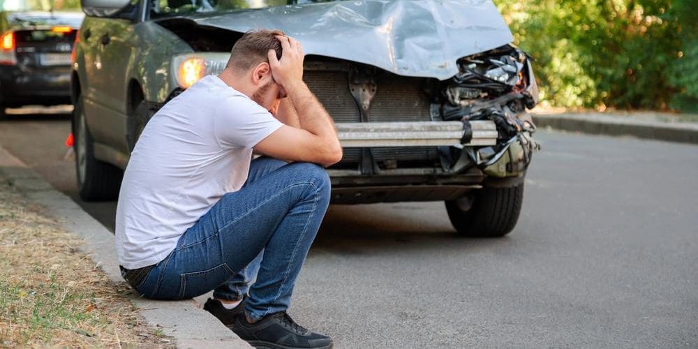 A man is sitting on the curb in front of his totaled car. He is suffering from a headache post accident.