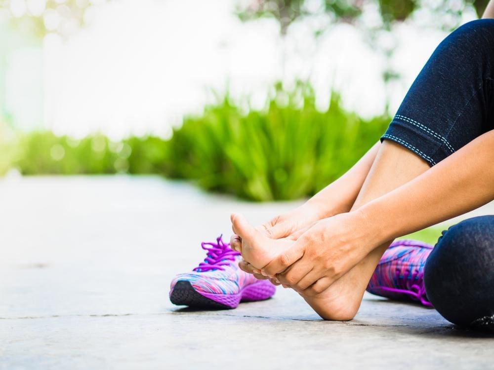 A woman is sitting on a sidewalk massaging her foot after removing purple running shoe.