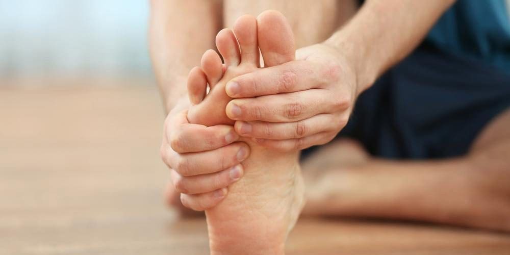 A middle-aged man is sitting on wooden chair clasping foot and grimacing because he is suffering from Morton's Neuroma.