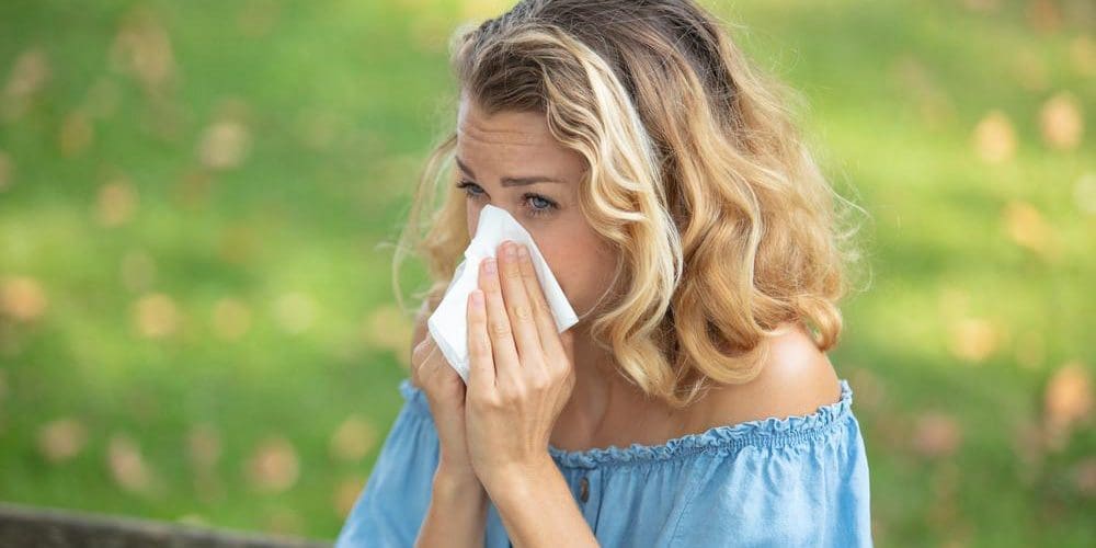 A woman is sneezing and blowing her nose while standing outside in a nice grassy field with flowers.