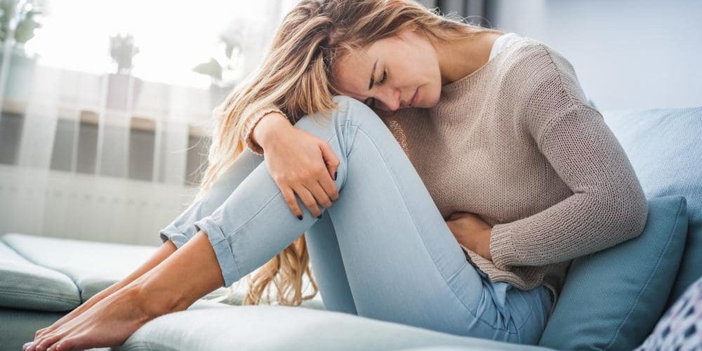 A woman is experiencing pms pain and hunching over on a couch clutching her abdomen.