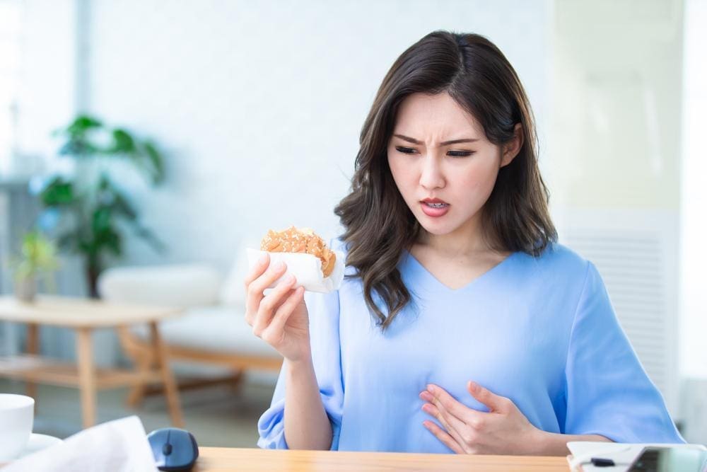 Woman eating a hamburger at her desk experiencing acid reflux.

