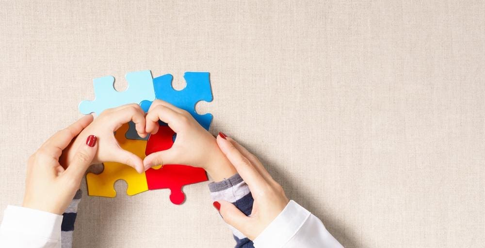 Woman holding child's hands in shape of heart of autism awareness puzzle pieces.

