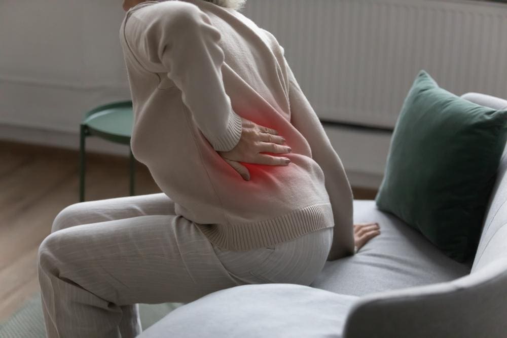 A woman is struggling to get up from the couch because of lumbar pain.