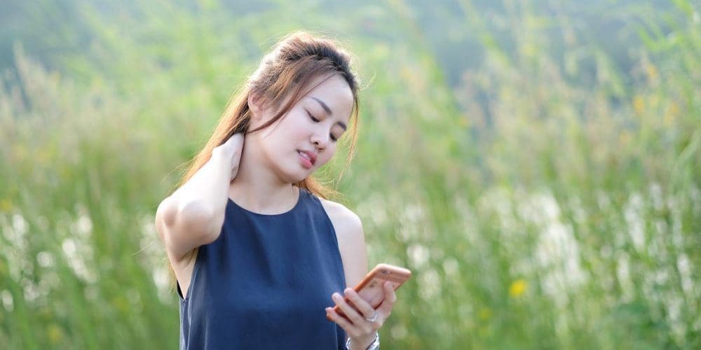 Young woman holding her neck while looking at her phone outside.