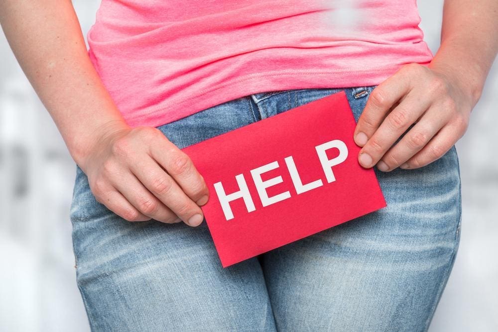 Closeup of a woman's hips in jeans and a t shirt holding a sign that reads "HELP" over her bladder.