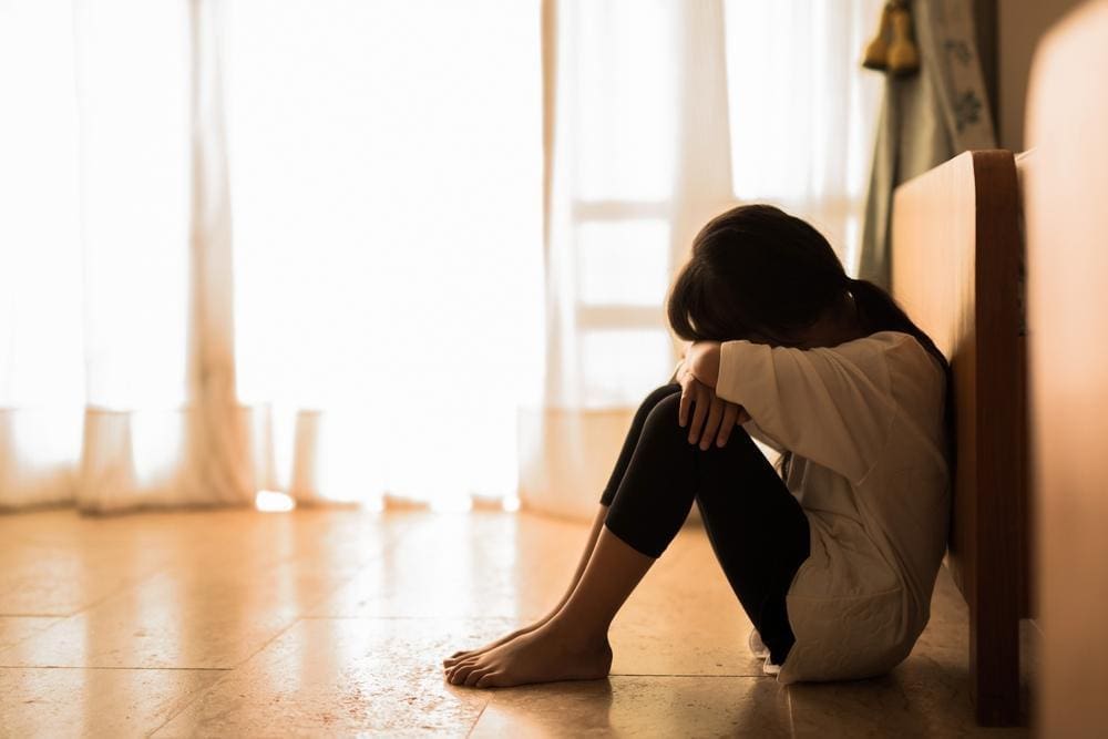 A young girl sits on the floor against the wall with her head in hands.