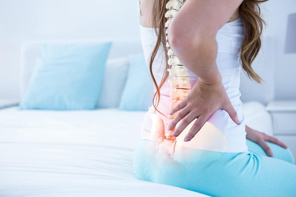A young woman sits on the edge of a bed with lower back pain.
