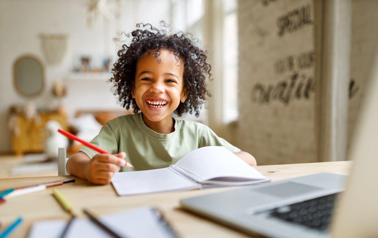 Smiling young boy doing schoolwork at a table in front of a laptop.