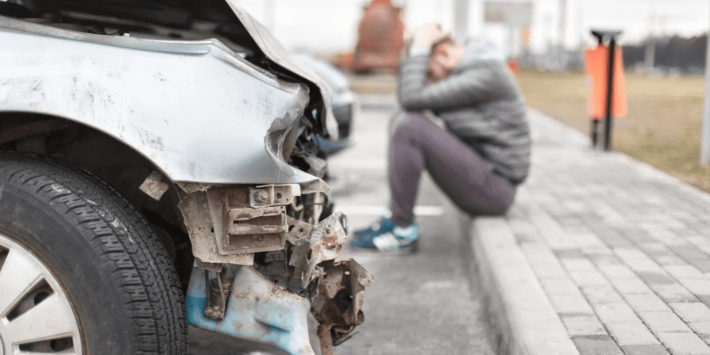 A man sitting on the curb next to his wrecked car.