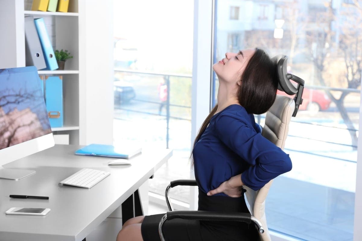 A woman leans back in her chair at work trying to alleviate lower back pain.