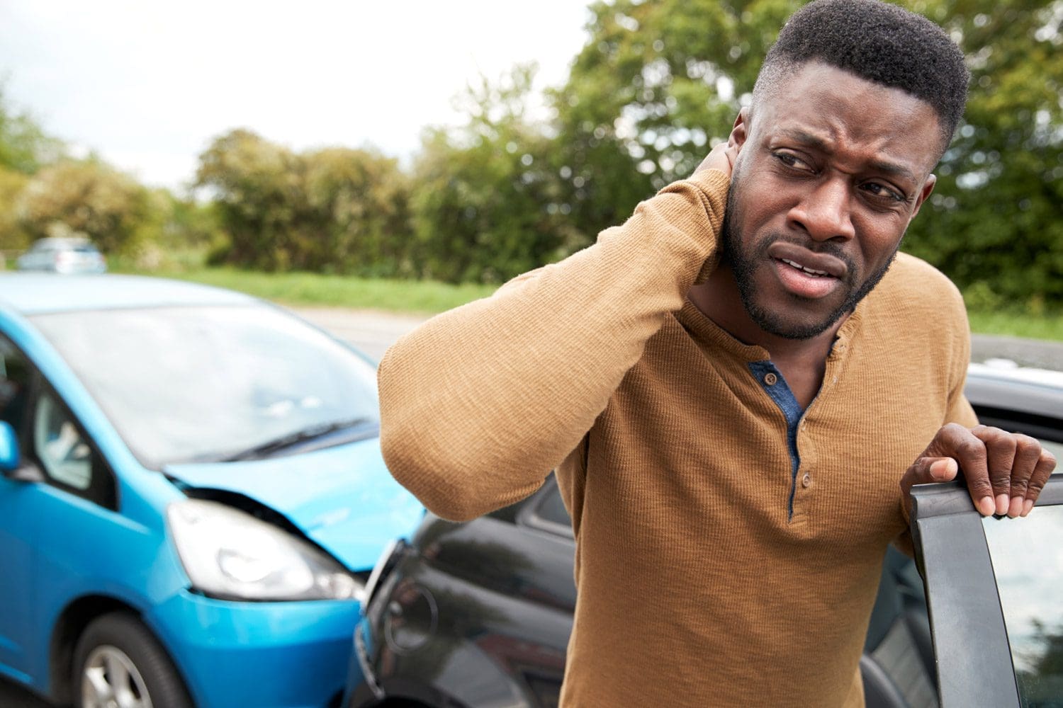 A man gets out of his wrecked car wincing in pain because of car accident injuries to his neck.