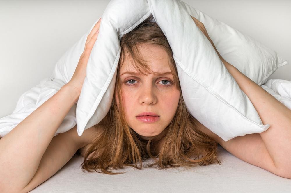 A young woman lying in bed frustrated holding pillow over her head, because she can't sleep. 
