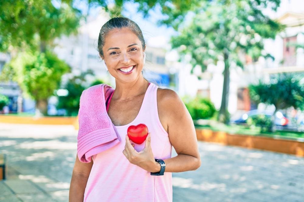 A happy and healthy middle-aged woman in athletic clothing holding red plastic heart to her chest.
