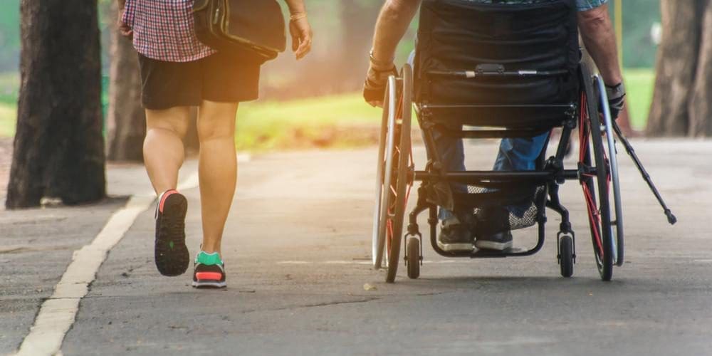 Woman walking alongside man in wheelchair holding cane.