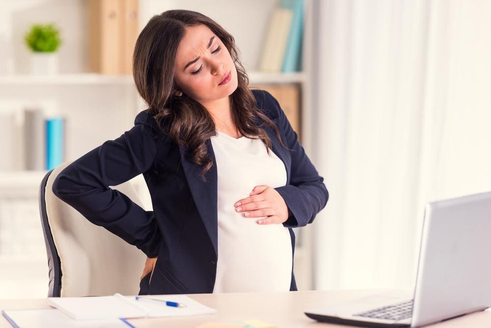Pregnant businesswoman holding her lower back and stomach while sitting in office chair.