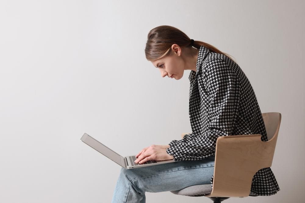 A young woman is hunched over in a chair looking down at a laptop on her lap.