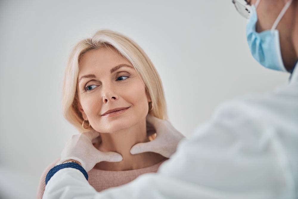 Healthcare provider wearing gloves and a mask assesses a woman's thyroid.