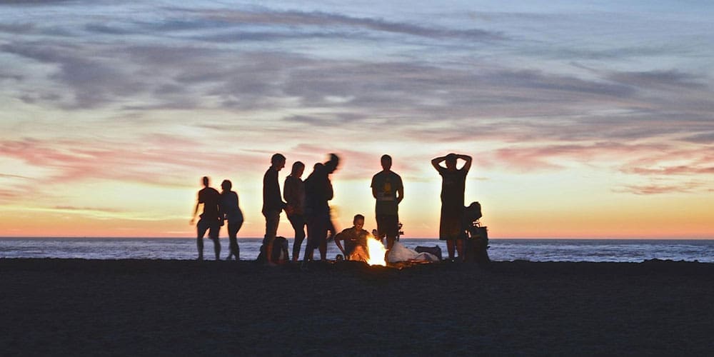 group of young adults around a fire on the beach at sunset